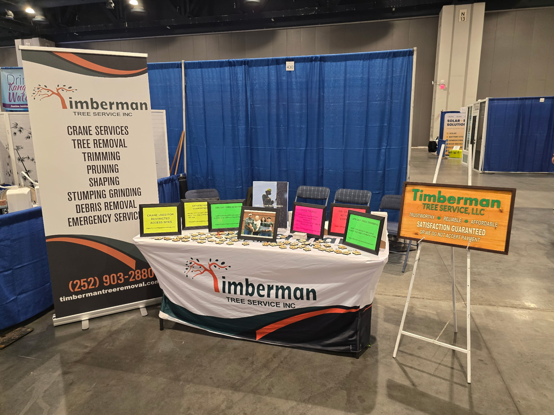 Timberman Tree Service booth at a Raleigh, NC home and garden show, with a branded table, service banner, and satisfaction guarantee sign